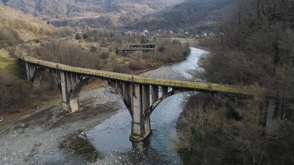 Aerial of abandoned concrete bridge covered by moss over the narrow river. Shot. Autumn landscape with high forested mountains and the small village.
