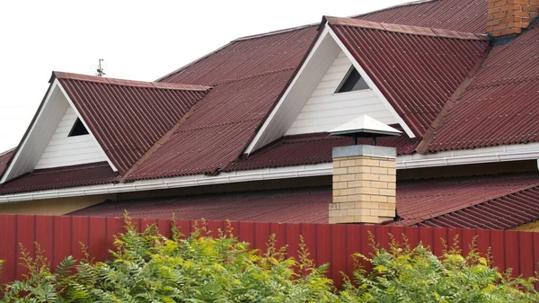 Σύγχρονη στέγη που καλύπτεται με πλακάκια του κόκκινου χρώματος σε γκρι φόντο του ουρανού, στέγες υλικά και την κατασκευή του κτιρίου έννοια. Στικ. Μοντέρνα πρόσοψη σπιτιού. — Φωτογραφία Αρχείου