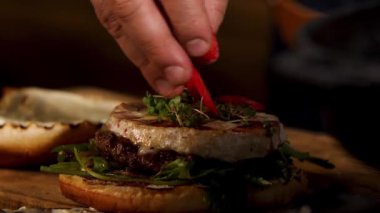 Delicious juicy red hot chili pepper burger with beef cutlet and camembert cheese, foodporn. Stock footage. Close up of male hand putting red circles of chili pepper in the burger.
