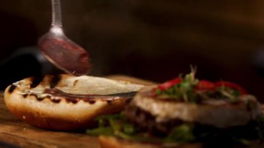 Male chef hands preparing burger in dark room and adding liquid red wine sauce on a toasted burger bun with a teaspoon. Stock footage. Foodporn and gastronomy concept.