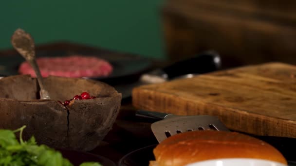 Close up of the ingredients on the wooden table. Stock footage. Beef cutlet, cranberries in wooden bowl, greenery, and burger bun near wooden board. — 비디오