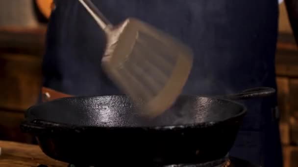 Close up of male chef frying ingredients in black iron pan and stirring them with a metal shovel. Stock footage. Process of food preparation. — 图库视频影像