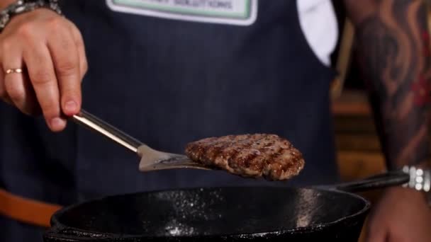 Dekat dengan koki laki-laki menggoreng daging burger di wajan hitam besi dari kedua sisi dengan bantuan sekop. Rekaman saham. Man in apron mempersiapkan daging potong . — Stok Video