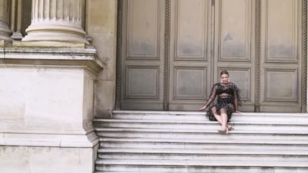 Atractiva mujer sexy sentada en las escaleras del edificio histórico y posando frente a la cámara. Acción. Rubia joven hembra en vestido provocativo negro . — Vídeos de Stock