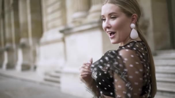 Schöne junge Frau in einem schwarzen Chiffonkleid, das provokant aussieht und lacht. Handeln. Sexy blondes Model lächelt vor altem Gebäude. — Stockvideo