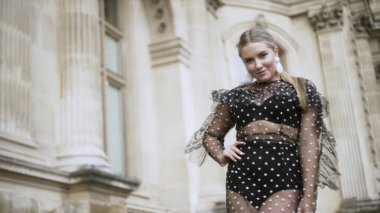 Genç ve cüsseli seksi manken, beyaz noktalı transparan siyah elbiseyi giyip antik katedralin önünde poz veriyor. Başla. Vücut pozitif konsept.