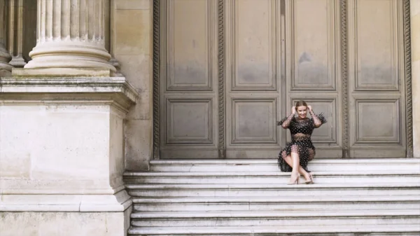 Atractiva mujer sexy sentada en las escaleras del edificio histórico y posando frente a la cámara. Acción. Rubia joven hembra en vestido provocativo negro . —  Fotos de Stock