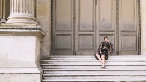 Atraktivní sexy žena sedící na schodech historické budovy a pózující před kamerou. Akce. Blond mladá fena v černošky provokativní šaty. — Stock fotografie