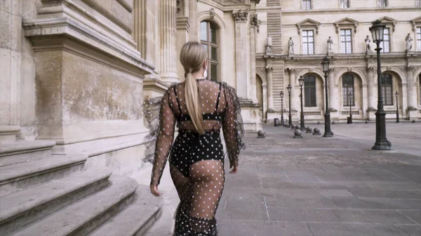 Jeune femme portant une robe noire transparente sexy tout en marchant dans la rue dans le centre historique de la ville européenne. L'action. Sexy attrayant modèle blond plus la taille en robe de mousseline de soie, vue arrière . — Photo