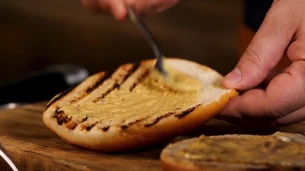 Molho de mostarda e mel para hambúrguer caseiro na cozinha. Imagens de stock. Close up de mãos cozinheiras masculinas adicionando mostarda e molho de creme azedo branco em um pão de hambúrguer . — Vídeo de Stock