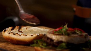 Male chef hands preparing burger in dark room and adding liquid red wine sauce on a toasted burger bun with a teaspoon. Stock footage. Foodporn and gastronomy concept.