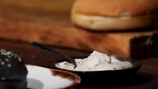 Close up of white sauce in black bowl standing on the wooden table near black and white burger buns, food ingredients concept. Stock footage. Home made mayonnaise sauce. — Stockvideo