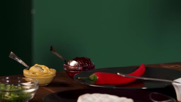 Close up of different ingredients for cooking standing on a wooden table on green wall background. Stock foorage. Chili pepper, cranberry, mayonnaise, and mustard sauces, camembert cheese and greenery — ストック動画