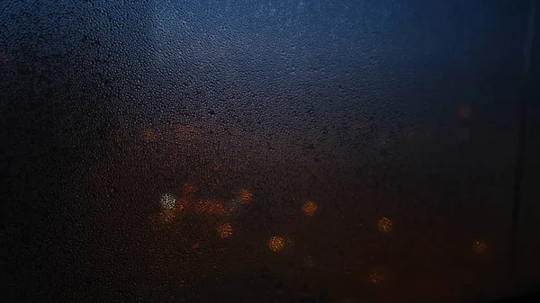 Egy közeli ablak esőcseppekkel a homályos fények hátterén. Fogantatás. Az esőcseppek által elmosódott sötét ablakon át halvány fények ragyognak. — Stock Fotó