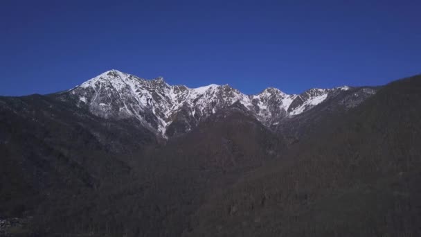 Vista panorámica de las montañas cubiertas de nieve sobre fondo azul claro del cielo. Clip. Paisaje aéreo de invierno con altas rocas con árboles en crecimiento en las laderas . — Vídeo de stock