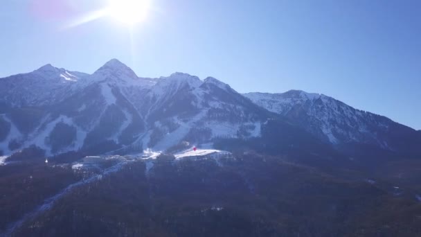 Vista aérea de la estación de esquí durante un vibrante día de invierno. Clip. Volando sobre las montañas nevadas boscosas en el cielo azul claro con sol brillante . — Vídeos de Stock