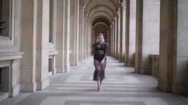 Young pretty woman with blond hair in black sexy dress and underwear walking near european historic building. Action. Attractive plus size model posing in front of the camera, body positive concept.