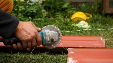 Roofer, çatı kiremitini kırmızı renkte kesmek için bir açı öğütücü makinesi kullanıyor. Stok görüntüleri. Erkek işçi elleri profesyonel testereyle çatı kiremiti keserken yakın plan..
