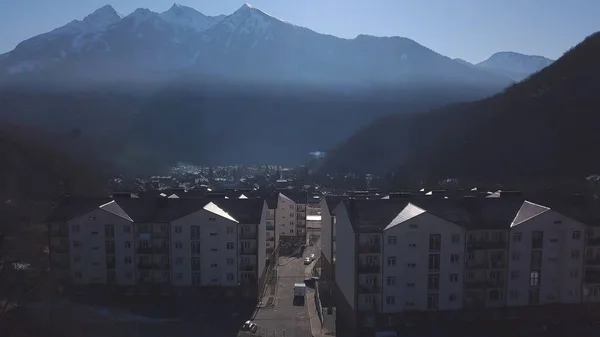 Aerial view of the small town situated in the valley surrounded by high mountains covered by trees. Clip. Flying above the town near forested hills on blue sky background. — ストック写真