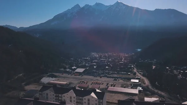 Aerial view of the small town situated in the valley surrounded by high mountains covered by trees. Clip. Flying above the town near forested hills on blue sky background. — ストック写真