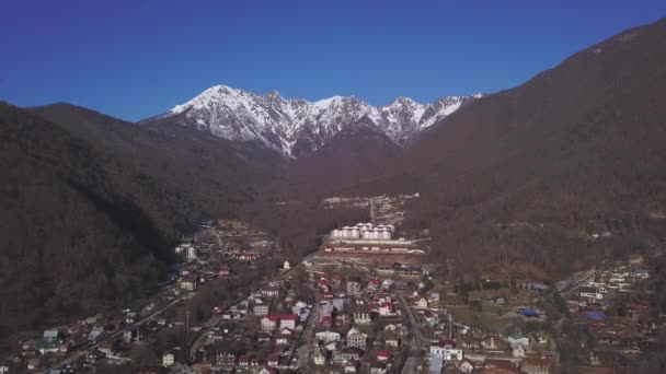 Aerial view of small village among the the trees located at the foot of the mountains in winter season. Clip. Winter picturesque landscape with snow covered mountain tops and the countryside. — 图库视频影像