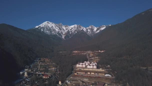Aerial view of small village among the the trees located at the foot of the mountains in winter season. Clip. Winter picturesque landscape with snow covered mountain tops and the countryside. — Stok video
