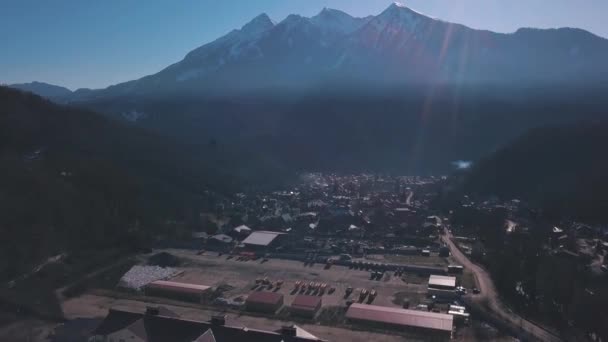 Aerial view of the small town situated in the valley surrounded by high mountains covered by trees. Clip. Flying above the town near forested hills on blue sky background. — Stockvideo