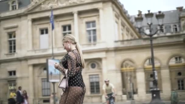 Boční pohled na neuvěřitelné blondýny v černých šatech chůze a pózování v blízkosti krásné historické budovy. Akce. Mladá žena s dlouhým rovným culíkem a bílou malou spojkou.