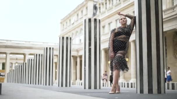 Chica de belleza con look glamour con vestido sexy transparente y ropa interior negra y de pie cerca de columnas a rayas en París, Francia. Acción. Mujer sensual con cabello rubio y look de moda . — Vídeo de stock