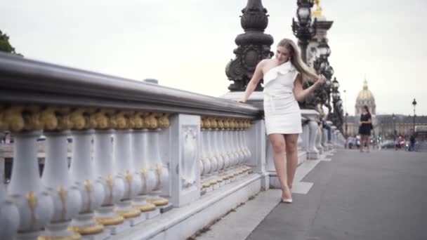 Prachtig blond meisje dat op de brug staat in witte stijlvolle jurk op de achtergrond van de grote stad. Actie. Sexy plus size model in korte jurk poseren op een mooie oude stenen brug. — Stockvideo
