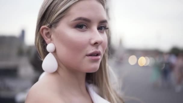 Portrait of a beautiful girl with blond hair standing outdoors on a bridge and admires the view of the big city. Action. Sensual woman face with big white earings smiling at the camera. — Stock Video