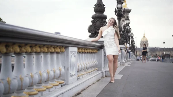 Lenyűgöző szőke lány áll a hídon fehér elegáns ruhát a nagyvárosi háttérben. Felvétel. Szexi plusz méret modell rövid ruhában pózol egy gyönyörű öreg kőhídon. — Stock Fotó