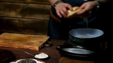 Erkek aşçı ellerinin hamburger ekmeğini alıp ahşap bir tahtanın üzerine koyması ve farklı yemek takımları ve malzemelerle çevrili olması. Stok görüntüleri. Lezzetli hamburger hazırlama süreci.