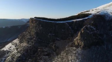 Yüksek kaya kütlesinin yamacı mavi gökyüzü arka planında çam ağaçlarıyla kaplıdır. Vuruldu. Ormandaki dağ yamacı ve karlı tepe..