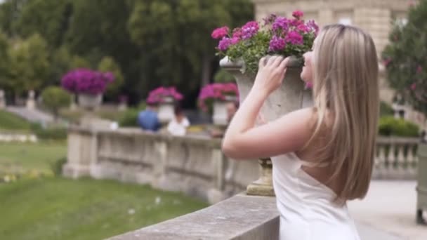 Loira em um vestido sexy branco no parque verde verão tocando seu cabelo longo com canteiros de flores roxas no fundo. Acção. Mulher atraente de pé em um jardim da cidade . — Vídeo de Stock