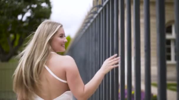 夏の日に格子鉄フェンスの近くを歩く白い柔らかいドレスの若い美しいブロンドの女性。行動だ。フェンスに沿って歩くプラスサイズモデルのリアビュー. — ストック動画