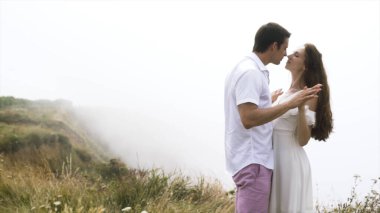Genç çift, arka planda kalın bir sis ile uçurumun kenarında sarılıyor. Başla. Şefkatli bir adam ve kadın dağdaki bulutların üstünde eğleniyorlar..