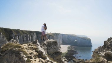 Kayaların üzerinde duran, sarılıp öpüşen, mutlu görünen, mavi gökyüzünde, denizde ve arka planda beyaz kayalıklarda aşık bir çift. Başla. Deniz kıyısının nefes kesici manzarası.