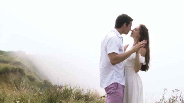 Pareja joven abrazándose en el borde del acantilado con espesa niebla en el fondo. Acción. Tierno hombre y mujer disfrutando unos de otros por encima de las nubes en una montaña . — Vídeos de Stock