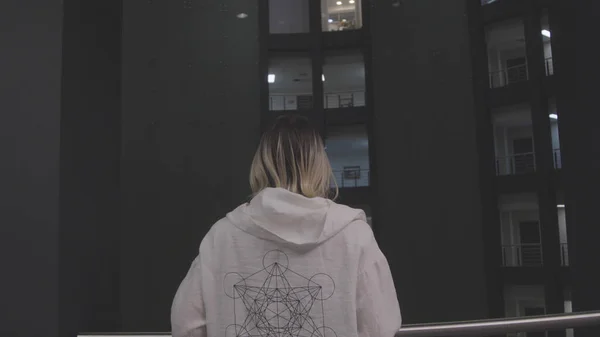 Woman looking down high rise. Stock. Back view of woman in hoodie looking down over railing of residential complex floor. Multi-storey residential apartment complex