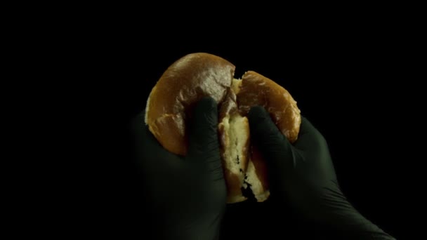 Close up de mão em luvas de cozinha pretas quebrando pão em dois pedaços isolados em fundo preto. Imagens de stock. Conceito de comida e culinária, pão de hambúrguer fresco sendo dividido . — Vídeo de Stock