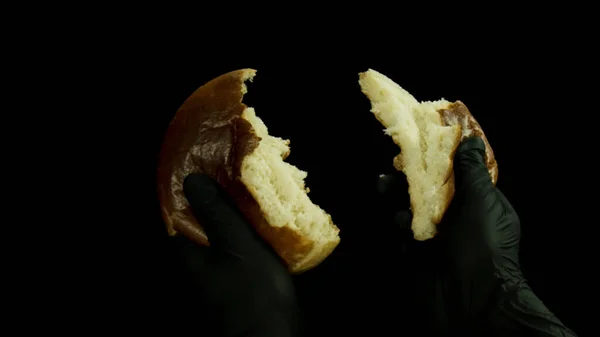 Close-up van de hand in zwarte kookhandschoenen breken brood in twee stukken geïsoleerd op zwarte achtergrond. Voorraadbeelden. Voeding en koken concept, verse hamburger broodje wordt gesplitst. — Stockfoto