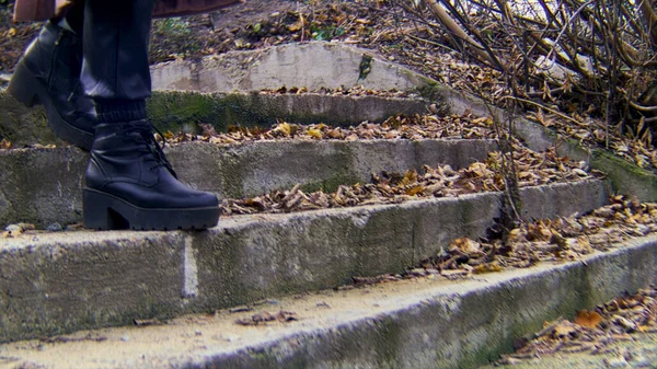 Botas femininas descendo as escadas em um dia de queda fria no parque da cidade. Imagens de stock. Close up de sapatos de couro preto descendo as escadas de granito com folhas de outono murchas . — Fotografia de Stock