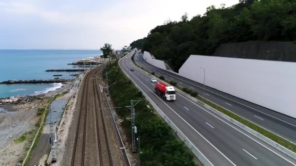 Aerial view of a truck driving on the road along the sea coast and rails, transportation concept. Scene. Cars moving on the road near the slope covered by green trees. — Stock Video