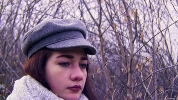 Νεαρή γυναίκα πορτρέτο με μια πολύ λυπητερή έκφραση στο φόντο φαλακρών φθινοπωρινών δέντρων. Στικ. Κοντινό πλάνο μιας σκεπτικής κοπέλας που φοράει ζεστό καπέλο και μαντήλι.. — Αρχείο Βίντεο