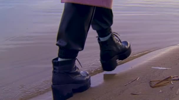 Primer plano de botas negras de cuero hembra caminando sobre una orilla arenosa y el borde del agua. Imágenes de archivo. Detalles de una joven caminando junto al río . — Vídeos de Stock