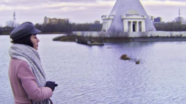 Jeune femme pensive en manteau rose debout au bord du lac regardant de côté, effet caméra VHS. Images d'archives. Jeune fille aux cheveux longs foncés debout près de l'eau en saison froide . — Video