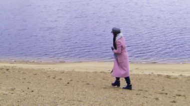 Sonbaharda gölün kenarında yalnız yürüyen pembe ceketli, esmer bir kadın, VHS etkisi. Stok görüntüleri. Yalnız kız soğuk havada kumda yürüyor..