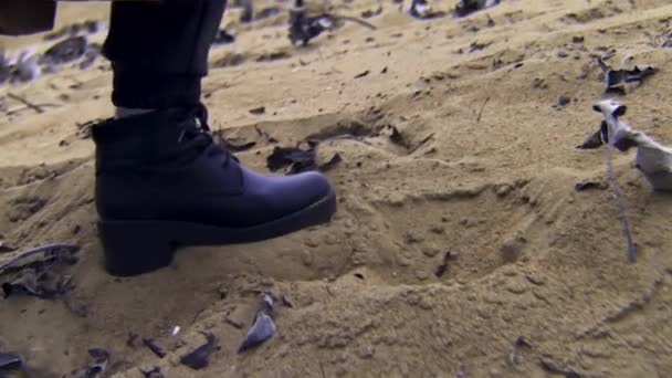 Primer plano de pies de mujer en botas negras caminando sobre arena con hojas secas. Imágenes de archivo. Mujer con botas de cuero negro caminando por la costa arenosa . — Vídeos de Stock