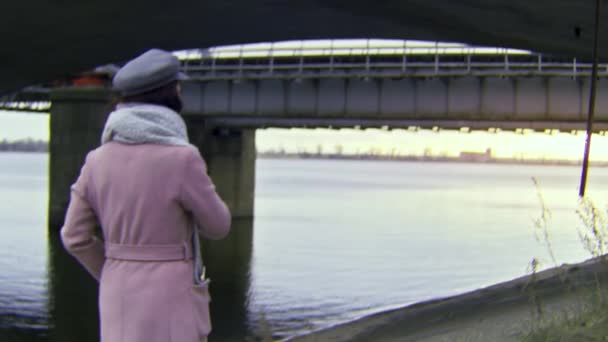 Achteraanzicht van een eenzame mooie vrouw in warme herfstkleding lopend onder de brug bij de rivier. Voorraadbeelden. Retro camera effect, romantisch meisje wandelen in een industriegebied. — Stockvideo